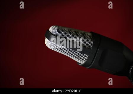 Close-up of a studio microphone against a dark red background, featuring a metal grille and black body, perfect for podcasts or music recording. Stock Photo