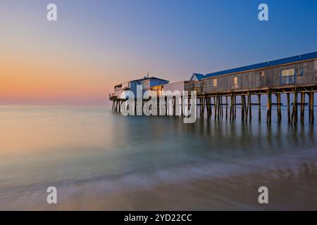 A serene ocean scene features a long wooden pier stretching into calm waters, with a soft gradient of hues in the sunset sky. The tranquil setting is Stock Photo