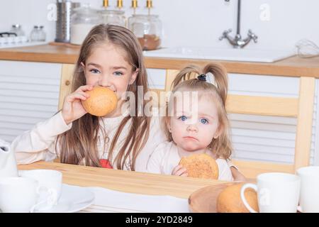 Two sisters at the table eating bread. Snack. Girls eat. Bright kitchen. Stock Photo