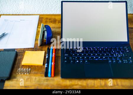 A laptop is on a desk with a stack of papers and a pen. The laptop is open and the screen is blank. The desk is cluttered with various items, includin Stock Photo