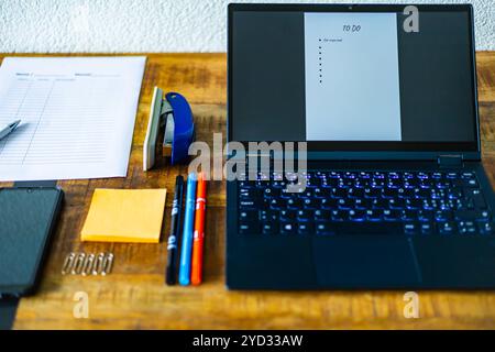 A laptop is open on a desk with a stack of papers and a pen. The laptop is displaying a list of tasks and a timer. The desk also has a stapler, a pair Stock Photo