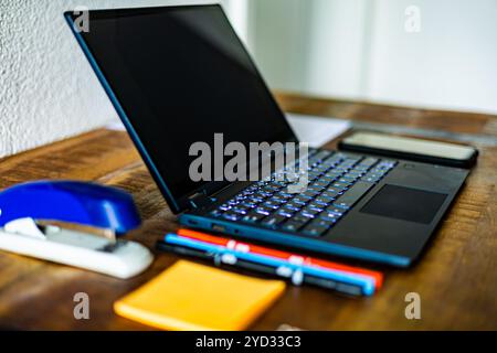 A laptop is open on a desk with a blue stapler, a red pen, and a yellow post-it note Stock Photo