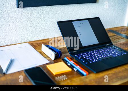 A laptop is open on a desk with a stack of papers and a pen. The laptop screen is displaying a list of tasks Stock Photo