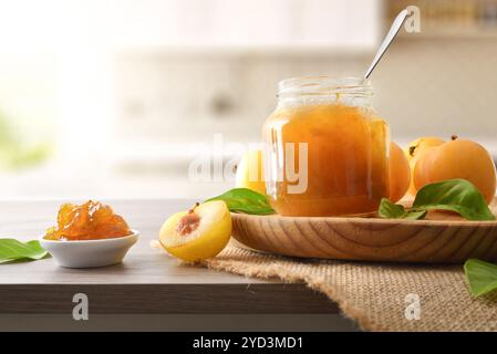 Jar of plum jam on wooden plate on bench and kitchen in background. Front view. Stock Photo