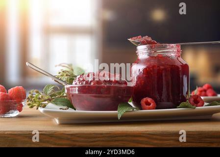 Detail of jars filled with raspberry jam on a white plate with leaves on a wooden table with bowl full of berries. Top elevated view. Stock Photo