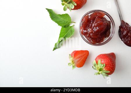 Glass bowl and spoon filled with strawberry jam isolated on white table with fruit around. Top view. Stock Photo