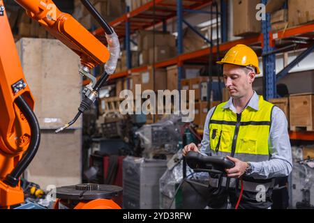 Engineer with safety gear using a control panel to operate an industrial robot in a manufacturing warehouse. Concept of modern technology, automation Stock Photo