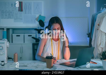 Young female fashion designer working late at night in her studio, focused on laptop and pattern sketches. Creative workspace environment. Stock Photo