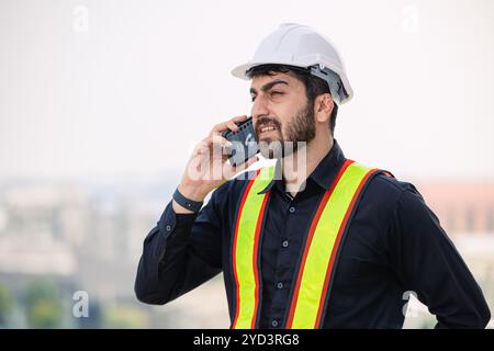 A construction worker wearing a safety helmet and vest speaks on the phone, emphasizing communication and safety at a worksite. Stock Photo