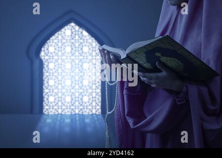 Religious muslim woman with hijab holding prayer beads while reading holy book of Al Quran in the mosque. Muslim holy book. Concept of Islamic religio Stock Photo