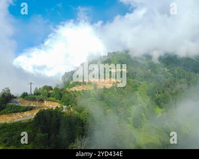 Winding mountain road disappears into clouds, with lush green forest below and blue sky peeking through, creating a mysterious and captivating scene Stock Photo