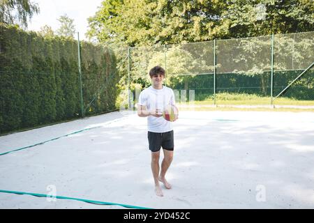 Young man holding a volleyball on a sand court, ready to play under a bright summer sky. Casual sports scene in a relaxed outdoor setting, perfect for Stock Photo