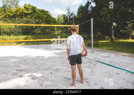 Volleyball player standing on a sand court, holding a ball and preparing for the next play during a quiet moment in an outdoor beach volleyball match. Stock Photo