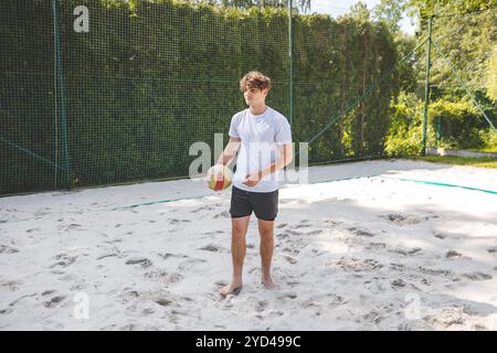 Volleyball player standing on a sand court, holding a ball and preparing for the next play during a quiet moment in an outdoor beach volleyball match. Stock Photo