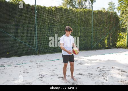 Volleyball player standing on a sand court, holding a ball and preparing for the next play during a quiet moment in an outdoor beach volleyball match. Stock Photo