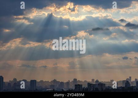 Dawn sun rays break through thick clouds over the silhouettes of houses in a sleeping city. Bright light in a dramatic morning sky. Stock Photo