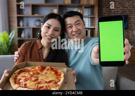 Asian couple shares joyous moment with pizza and smartphone in cozy living room. Man holds phone displaying green screen, adding playful twist to their happy delivery meal experience. Stock Photo