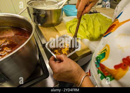 An employee of the Le Sfogline pasta workshop ladles meat ragù from a pot into a foil tray, while a green sheet of pasta dough and a pot of pale sauce wait in the background for preparing the green lasagne. Via Belvedere, Bologna, Emilia-Romagna, Italy. Stock Photo