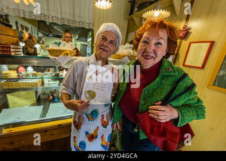 An employee of the Le Sfogline pasta workshop shows a customer a handwritten card illustrated with a leaf motif. In the background, a second employee is visible in the kitchen, reflected in the shop mirror. Via Belvedere, Bologna, Emilia-Romagna, Italy. Stock Photo