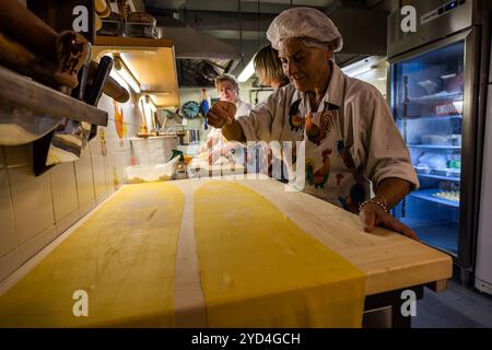 At the work table of the Le Sfogline pasta workshop, an employee stretches out a long strip of yellow pasta dough, while two colleagues prepare other ingredients in the background. Le Sfogline, at Via Belvedere 7/b, is run by the sisters Monica and Daniela, who took over the business from their mother, Renata Zappoli. Via Belvedere, Bologna, Emilia-Romagna, Italy. Stock Photo