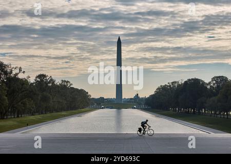 The early morning light casts a serene glow over the reflecting pool as a cyclist rides leisurely, with the iconic Washington Monument towering in the Stock Photo