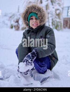 latino boy squatting in the snow in a fur hooded jacket, looking at the camera and holding a snowball. Stock Photo