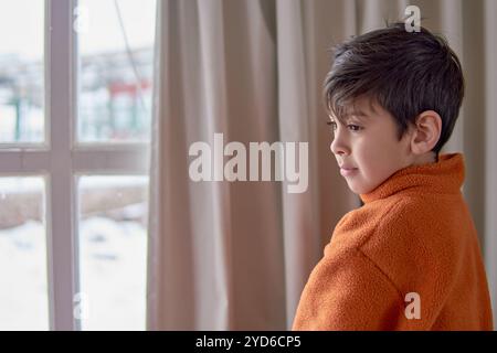 latino boy standing next to an open window with light curtain inside his house looking sideways pensively while it is snowing outside Stock Photo