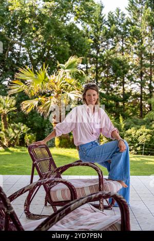 Woman sits on arm of chair in garden of luxury villa in South of France. Stock Photo