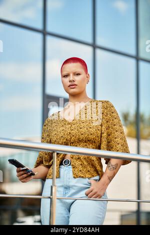 The beautiful bald woman poses confidently on a balcony, holding a phone against a bright sky. Stock Photo
