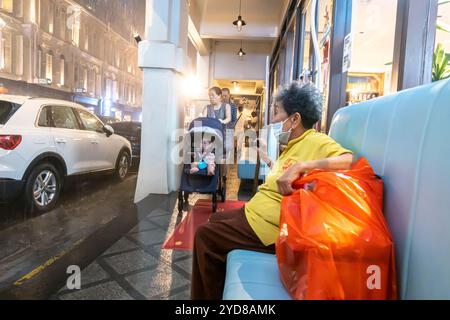 Singapore Chinatown,  Mosque street, rain, senior Chinese woman sitting on bench, couple with a baby stroller carriage Stock Photo