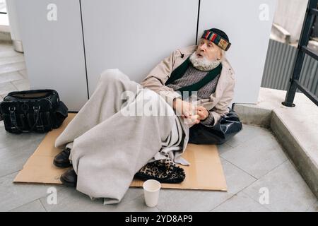 High-angle view of elderly homeless man with long white beard sitting lying on sidewalk with belongings, holding cup and seeking assistance. Stock Photo