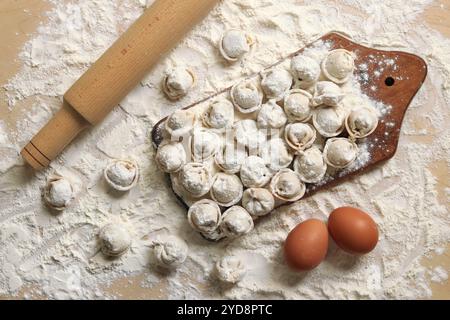 Uncooked dumplings, top view, flat lay. Cooking dumplings, flour, eggs and wooden rolling pin. Dumplings or ravioli on wooden board, table covered wit Stock Photo