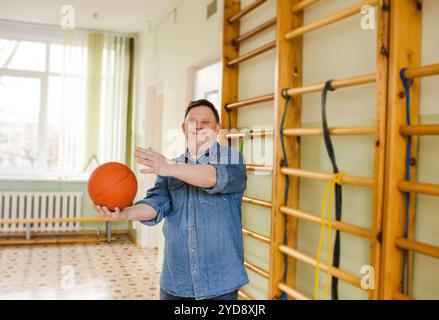 A young man with Down syndrome is playing basketball in a gym. He is smiling brightly, demonstrating the joy and benefits of physical activity for eve Stock Photo