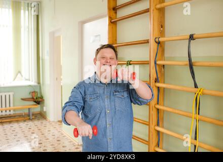 A man with Down syndrome is smiling and working out with dumbbells in a gymnasium. Stock Photo