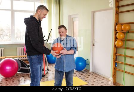 A man with Down syndrome is working out in a gymnasium with his trainer. They are using a medicine ball. Stock Photo