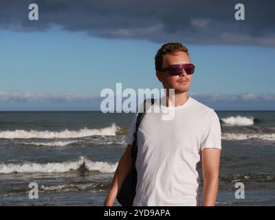 A young man with a backpack, wearing a white T-shirt and sunglasses standing on the seashore at the background of gloomy cloudy sky, with copy space. Stock Photo