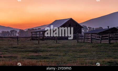 A large, old barn sits in a field with a beautiful sunset in the background. The scene is peaceful and serene, with the barn and the surrounding lands Stock Photo