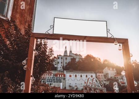 Outdoor wooden mockup frame with blank white sign, decorated with hanging string lights, against a sunset backdrop of a historic European cityscape wi Stock Photo