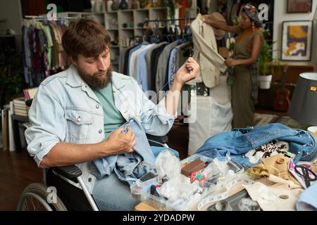 Young bearded male worker of modern thrift shop sitting in wheelchair by workplace and upcycling secondhand blue jeans Stock Photo