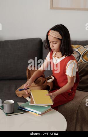 Girl in red dress concentrating on homework at home, sitting on couch surrounded by books and cup of coffee, creating a focused and calm atmosphere Stock Photo