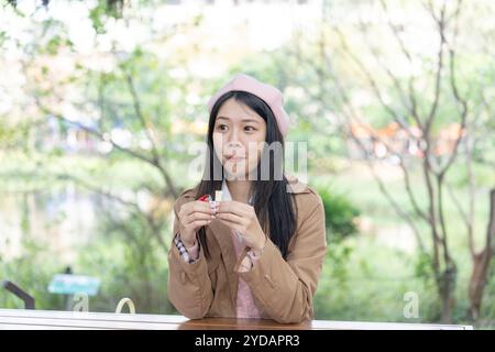 A long-haired Taiwanese woman in her 20s wearing a brown trench coat sits on a bench in a park in Xinyi District, Taipei, Taiwan and eats chocolate at Stock Photo