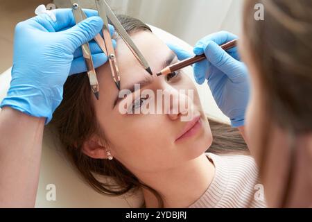 Changing the shape of the brows. Stylist measuring the eyebrows with the ruler. Micropigmentation work flow in a beauty salon. Woman having her eye br Stock Photo