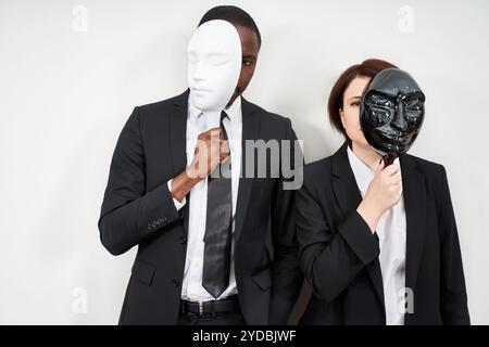 African man and Caucasian woman wearing black suits hiding face with plastic masks Stock Photo