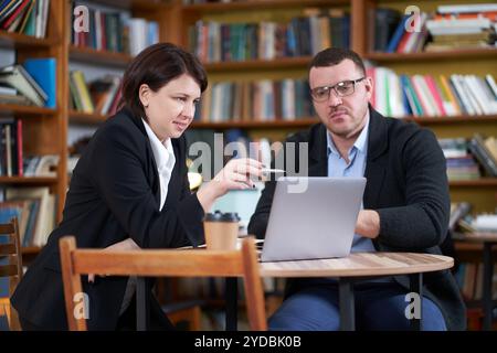 Man and woman working with laptop sitting in modern stylish room with bookshelves on background in library or bookstore Stock Photo