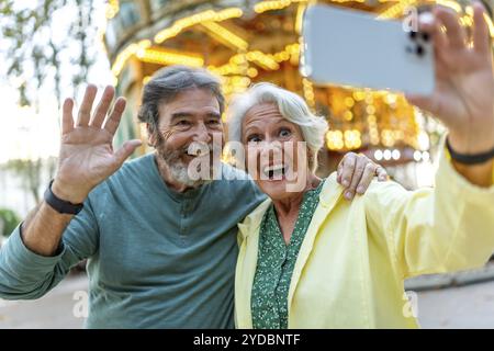 Senior couple waving at camera while taking selfie in the city next to carousel Stock Photo