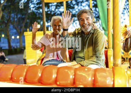 Senior happy caucasian couple waving sitting on a colorful carousel at night in the city fair Stock Photo