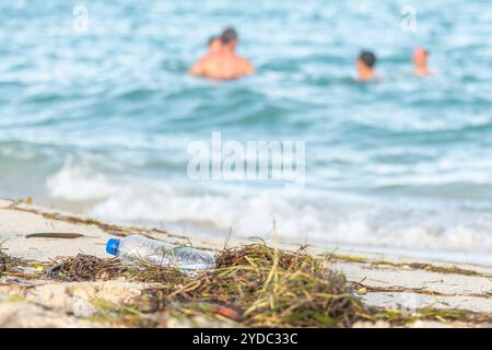 Close up image of empty plastic water bottle on dirty beach filled with seaweed, garbage and waste on dirty sandy beach with peo Stock Photo