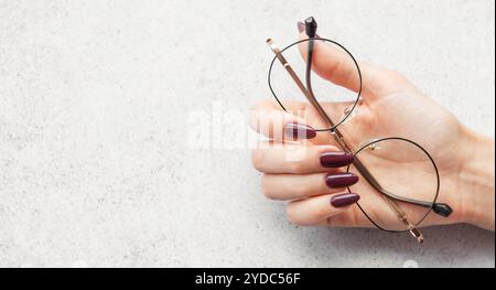 A hand with long burgundy nails gracefully holds a pair of round glasses above a light textured surface, illuminated by soft natural light. Stock Photo