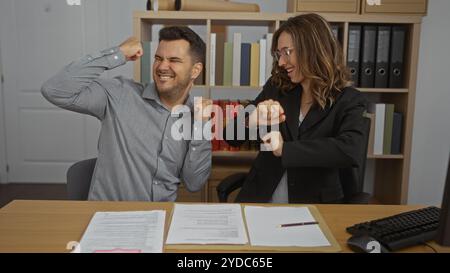 Man and woman celebrating in office with documents on desk and bookshelves in background, indicating a successful business deal or project completion Stock Photo