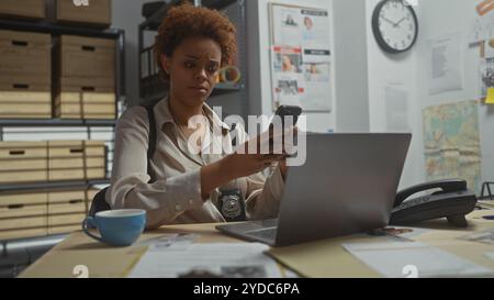 African american female detective using smartphone in police station office, surrounded by investigation evidence. Stock Photo